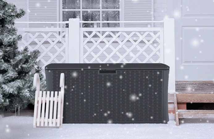 A gif of snow falling while a grey suncast deck box sits outside next to an evergreen tree and in front of a white fence.